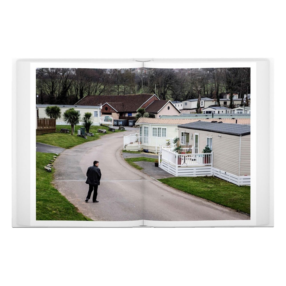 Spread shot of Jonas Bendiksen: The Last Testament, showing a full-width color photo of a man on the street in front of house.
