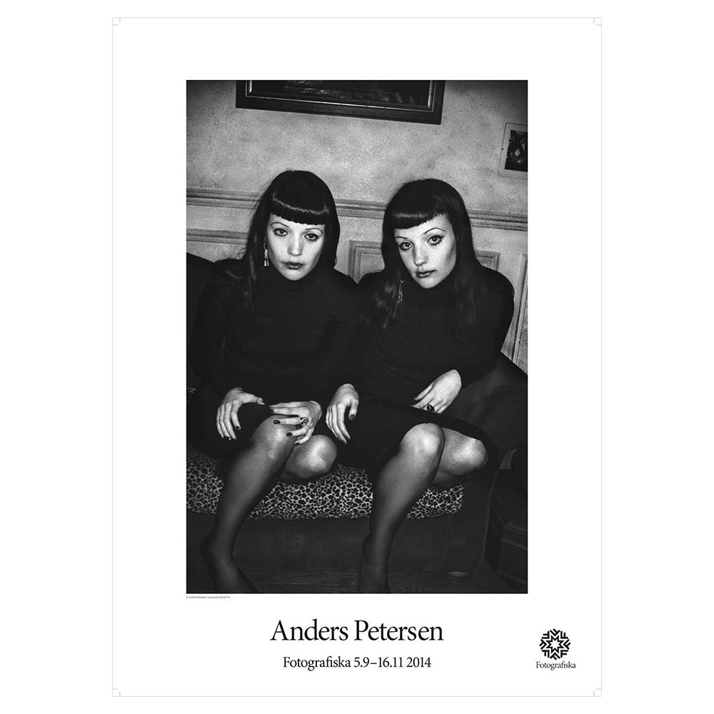 Black & white portrait of two identical female twins, dressed in black and sitting on a couch. Artist name below: Anders Petersen