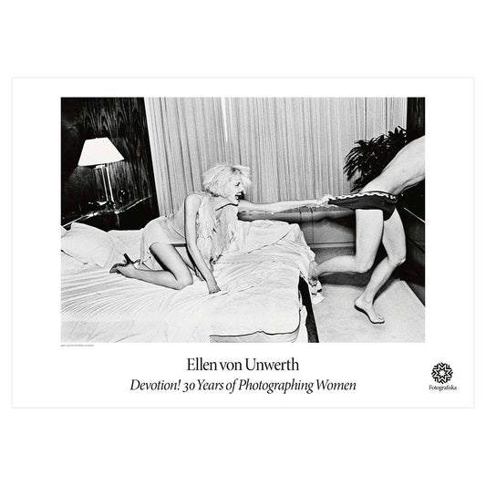 Black and white of a smiling woman on a bed pulling someone by their underwear as they are running. Exhibition title below: Ellen Von Unwerth: Devotion! 30 Years of Photographing Women